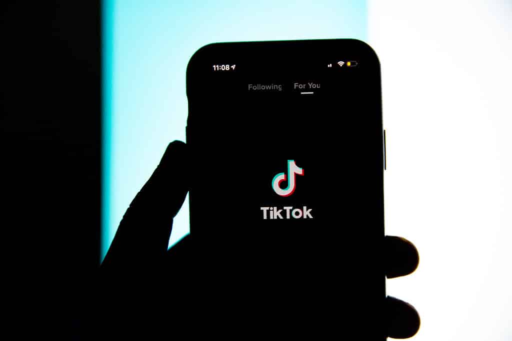 A hand holding a smartphone with the TikTok app open. The app's logo, a musical note icon, is displayed prominently in the center of the screen. The background is divided into dark and light blue vertical sections, creating a stark contrast with the silhouette of the hand and phone.