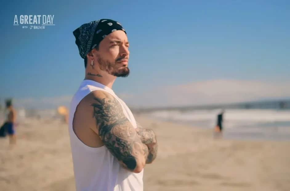 J Balvin, a man with tattoos, stands on a sunny beach, looking into the distance. Sporting a white sleeveless shirt and a black bandana, he exudes confidence. In the background, the ocean and a few people are visible. The text "A GREAT DAY with J Balvin" hints at his upcoming Peacock show in the top left corner.