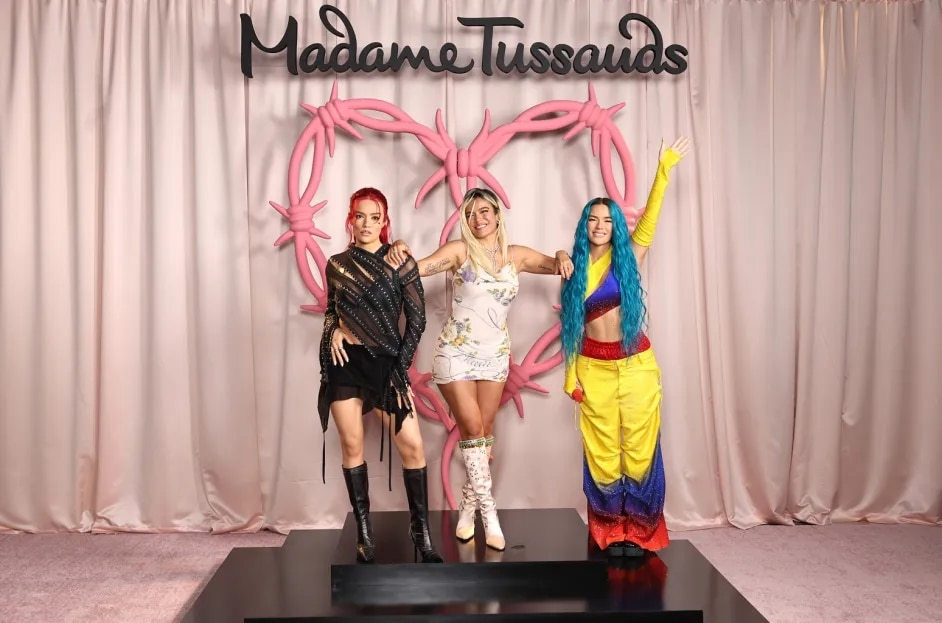 Karol-G and her Wax figures stand under a "Madame Tussauds" sign. The one on the left wears a black dress and boots, the center one in a white outfit, and the right one in a colorful ensemble with blue hair. They pose confidently on a stage with a pink, balloon-style heart backdrop.