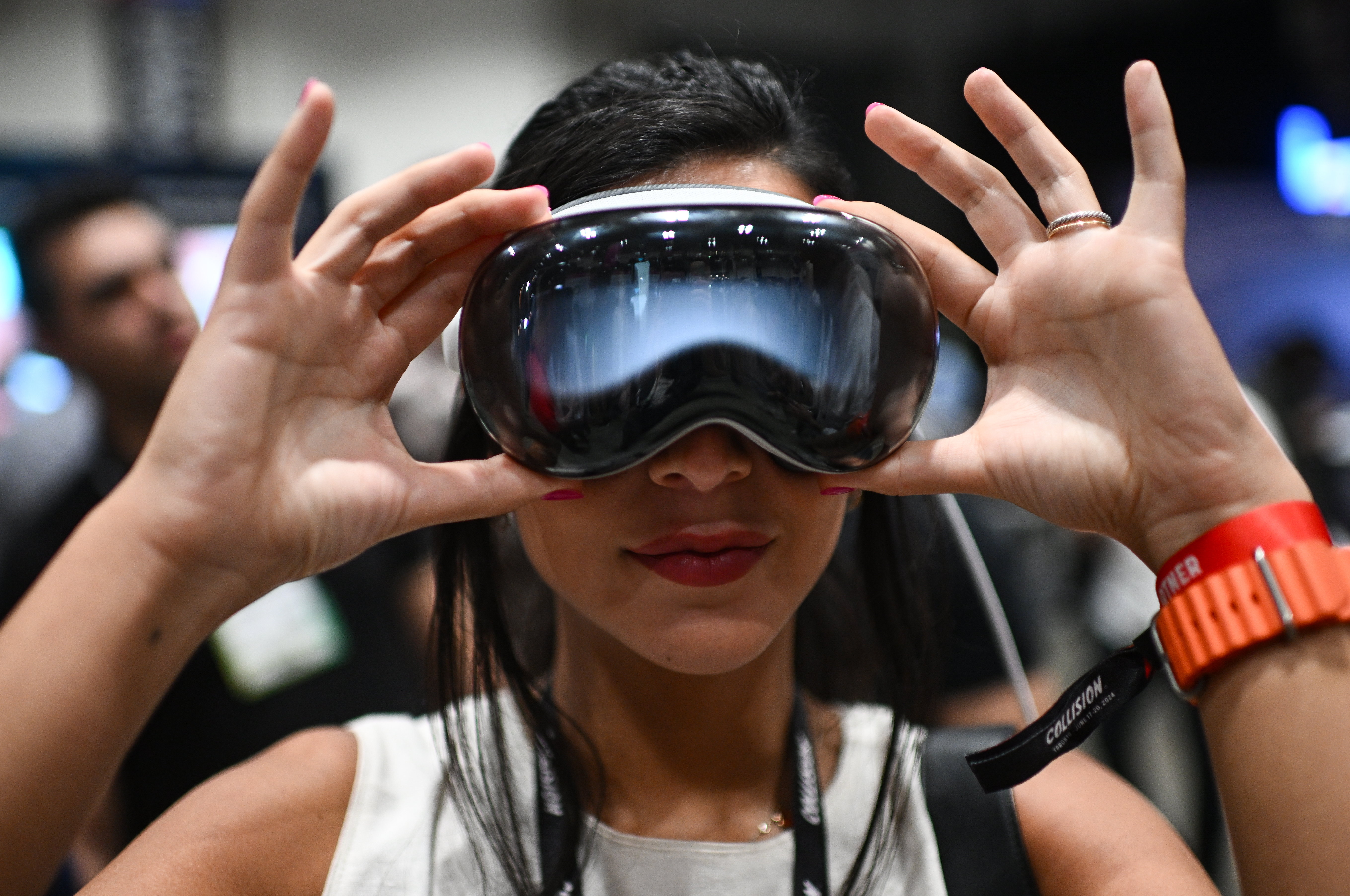 A person wears sleek, metallic virtual reality goggles, holding them with both hands. They have long dark hair and wear a sleeveless top. A red wristband is visible on one wrist. The blurred background hints at the future of virtual reality films and the Apple Immersive Video experience.