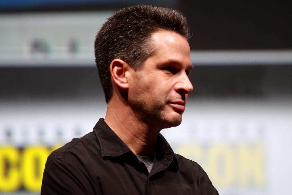 Man with short dark hair and a beard, Simon Kinberg, wearing a black shirt, standing in front of a blurred background featuring text and a horizontal screen. He is facing slightly to the right with a neutral expression. The setting appears to be an indoor event.