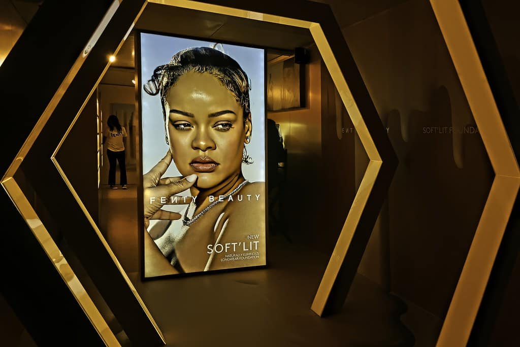 A digital display in a geometric, gold-toned hallway showcases an advertisement for Fenty Beauty's new Soft Blurring Primer, featuring a woman with a confident expression. She is elegantly styled, with a focus on flawless skin. Reflections and shadows add depth to the scene.