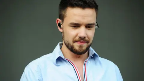 A man with short dark hair and a beard, Liam Payne, is looking down, wearing a light blue shirt with a red, white, and blue striped collar. He has an in-ear monitor in his left ear. The background is plain and dark, highlighting him in the foreground.