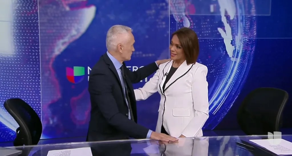 Jorge Ramos and Ilia Calderon, man and a woman are standing in a news studio. The man, in a dark suit, gently rests his hand on the woman's shoulder. She is wearing a white blazer with black trim. Behind them is a large digital screen displaying abstract graphics and partial text.