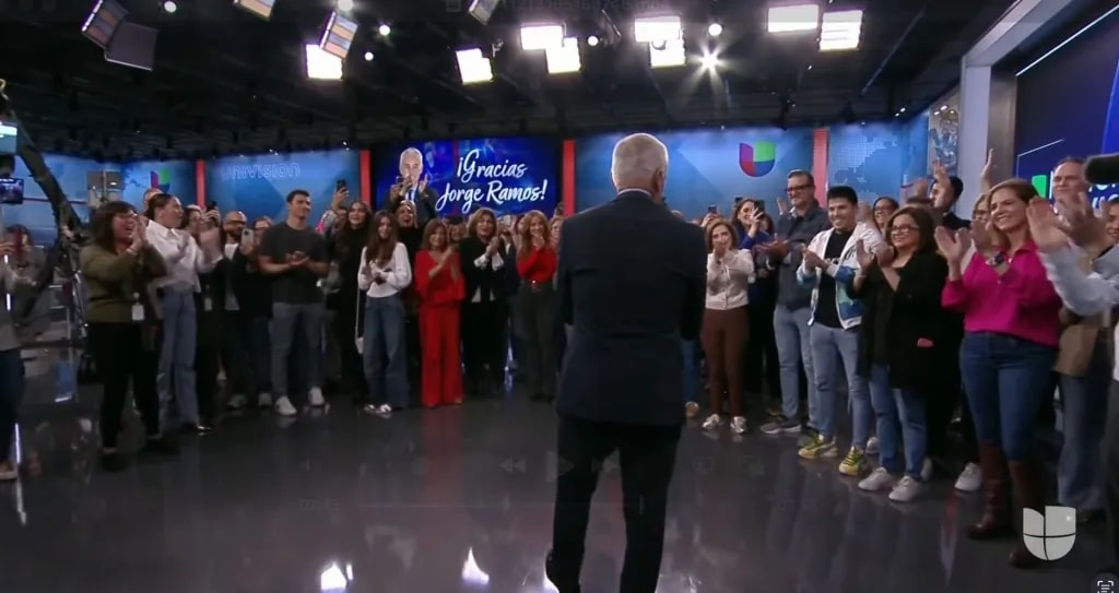 Jorge Ramos, a man stands with his back to the camera, facing a large crowd applauding in a TV studio. A backdrop reads "Gracias Jorge Ramos!" The crowd, casually dressed, consists of men and women smiling and clapping, while bright studio lights illuminate the scene.