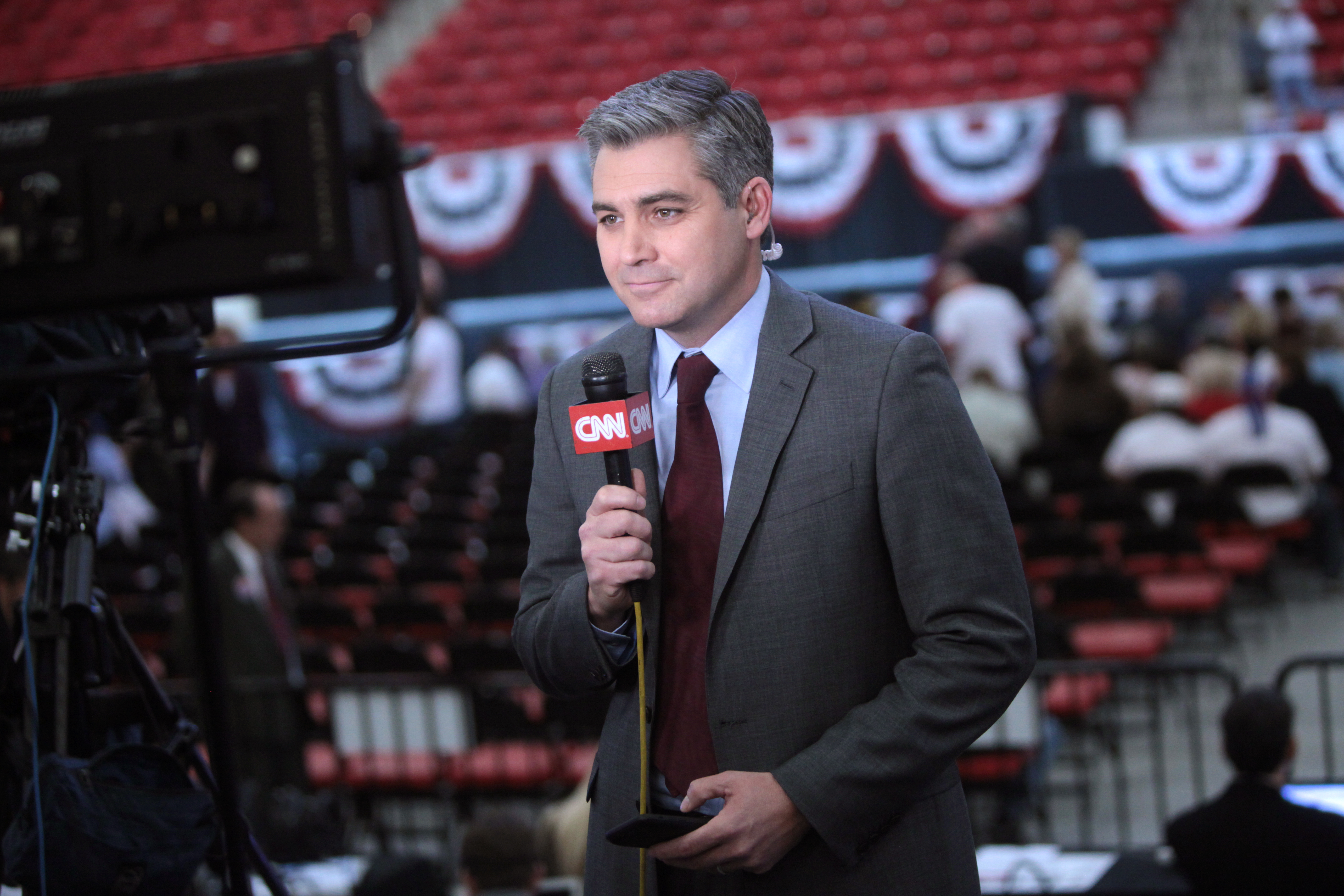 Jim Acosta, a man in a gray suit and maroon tie holds a microphone with a CNN logo, standing in an indoor venue. He appears to be reporting, with empty red seats and patriotic banners visible in the background. He has an earpiece in his left ear, and people are gathered in the distance.