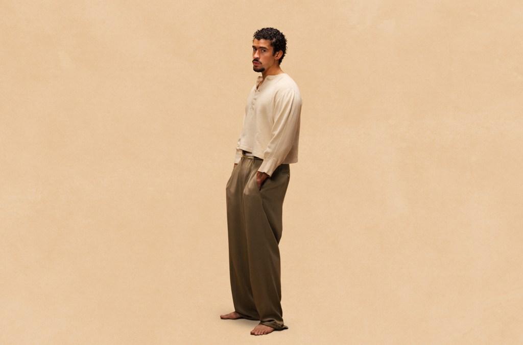 Bad Bunny, A barefoot man stands against a beige background, looking to the side. He wears a loose-fitting, long-sleeved, beige shirt and wide-legged, olive-green pants. His hands are in his pockets, and he has short, curly hair and a mustache.