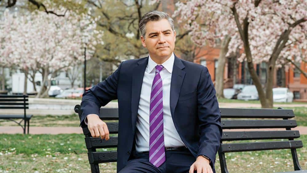 Jim Acosta, a man in a dark suit and purple striped tie sits on a park bench. He has short gray hair and is gazing to his left. The background features blooming trees and a building, suggesting a spring setting in an urban park.