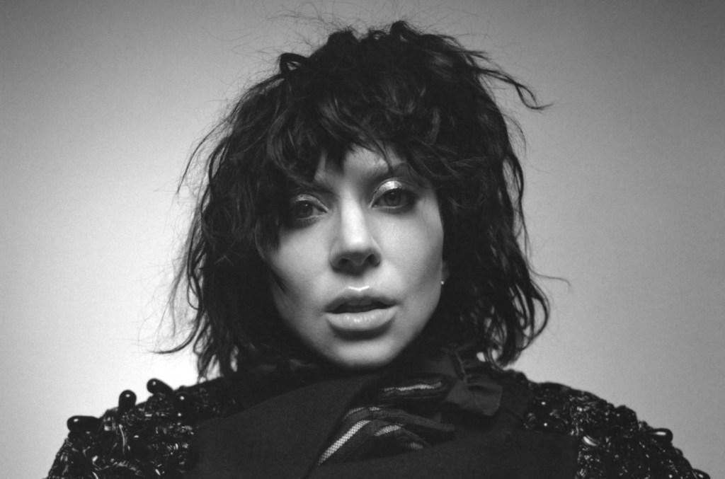 Black and white photo of a Lady Gaga with tousled, shoulder-length hair. They have a serious expression, wearing shimmery eye makeup, with lips slightly parted. The clothing has a textured, layered collar, adding depth to the somber tone of the image.