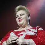 Paquita la del Barrio, a person with a confident expression, wearing a red sequined dress with white swirl patterns. They have short, styled hair and are adorned with dangly earrings and multiple rings. The background is a gradient of dark to light, suggesting a spotlight effect.