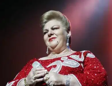 Paquita la del Barrio, a person with a confident expression, wearing a red sequined dress with white swirl patterns. They have short, styled hair and are adorned with dangly earrings and multiple rings. The background is a gradient of dark to light, suggesting a spotlight effect.