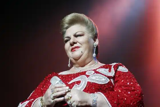 Paquita la del Barrio, a person with a confident expression, wearing a red sequined dress with white swirl patterns. They have short, styled hair and are adorned with dangly earrings and multiple rings. The background is a gradient of dark to light, suggesting a spotlight effect.