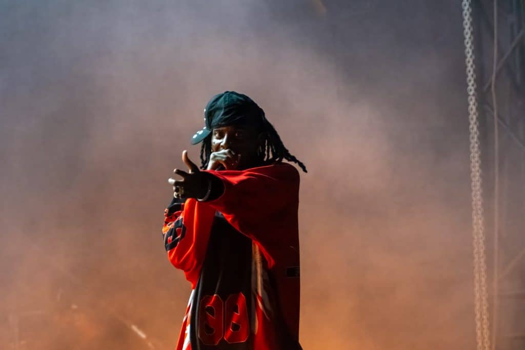 A performer on stage, wearing a red sports jersey and a black cap turned backward, points towards the audience while holding a microphone. Thick smoke envelops the stage, creating a dramatic atmosphere reminiscent of anticipation when Playboi Carti teases his 2024 album release.