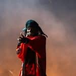 A performer on stage, wearing a red sports jersey and a black cap turned backward, points towards the audience while holding a microphone. Thick smoke envelops the stage, creating a dramatic atmosphere reminiscent of anticipation when Playboi Carti teases his 2024 album release.