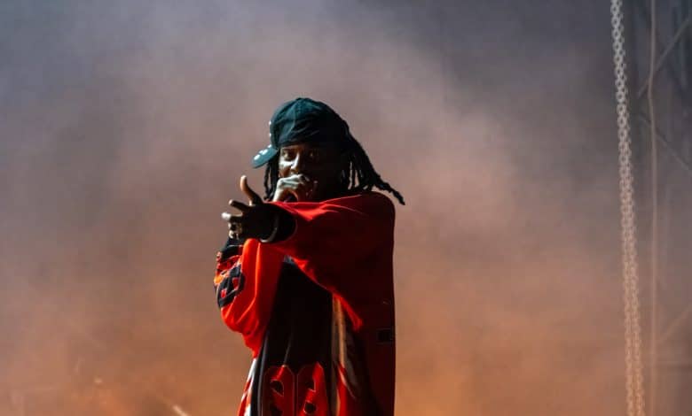 A performer on stage, wearing a red sports jersey and a black cap turned backward, points towards the audience while holding a microphone. Thick smoke envelops the stage, creating a dramatic atmosphere reminiscent of anticipation when Playboi Carti teases his 2024 album release.