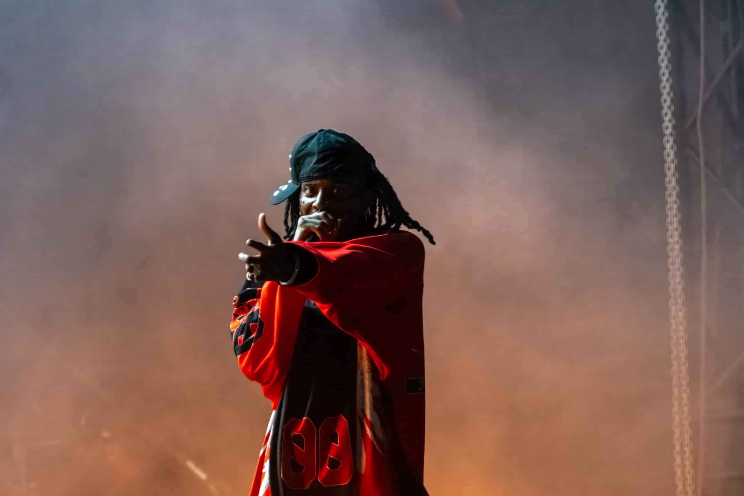 A performer on stage, wearing a red sports jersey and a black cap turned backward, points towards the audience while holding a microphone. Thick smoke envelops the stage, creating a dramatic atmosphere reminiscent of anticipation when Playboi Carti teases his 2024 album release.