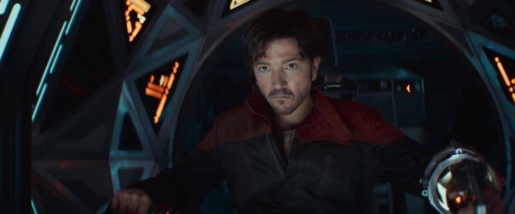 A man with short, dark hair sits in a spaceship cockpit, looking serious. He wears a dark jacket with a red collar. The cockpit is illuminated by soft, glowing lights with various control panels. He appears focused, with his hands positioned on the controls.