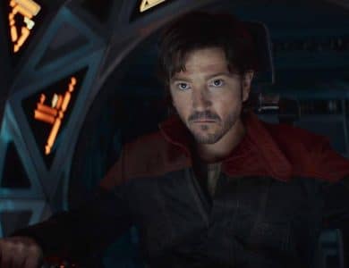 A man with short, dark hair sits in a spaceship cockpit, looking serious. He wears a dark jacket with a red collar. The cockpit is illuminated by soft, glowing lights with various control panels. He appears focused, with his hands positioned on the controls.