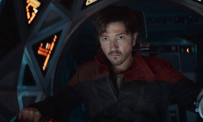 A man with short, dark hair sits in a spaceship cockpit, looking serious. He wears a dark jacket with a red collar. The cockpit is illuminated by soft, glowing lights with various control panels. He appears focused, with his hands positioned on the controls.