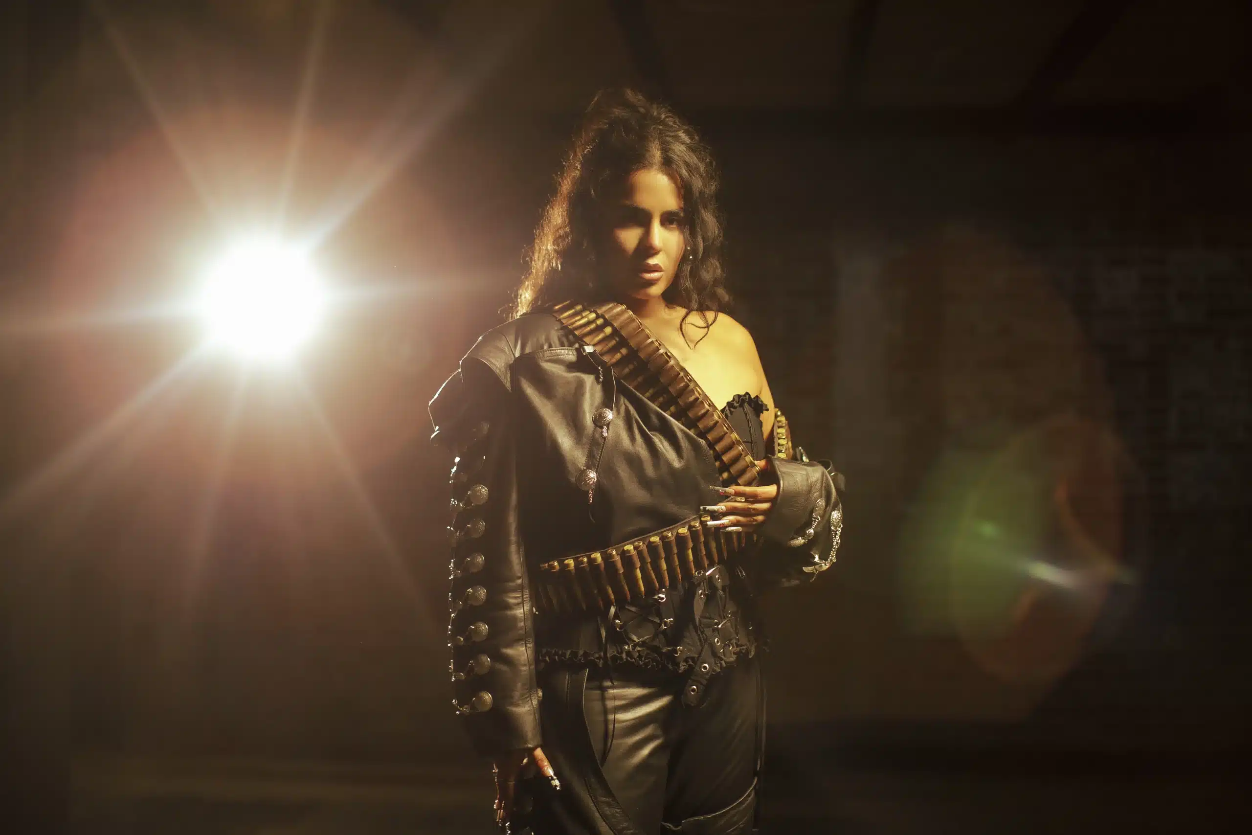 A woman stands confidently in dim lighting, channeling the energy of Immasoul LaDiosa as she poses with a bold expression. She wears a black, off-the-shoulder leather jacket adorned with bullet straps and matching pants. The dark background is pierced by a bright light creating a lens flare effect near her.