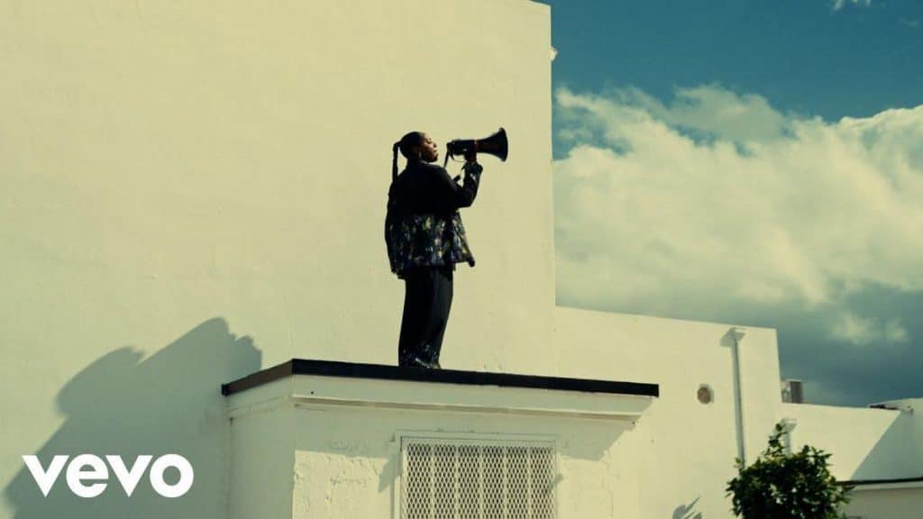 A person stands on a white rooftop holding a megaphone, speaking into it. They wear dark clothing and the sky behind is blue with scattered clouds. The building is modern and white; part of a leafy tree is visible below. The Vevo logo appears in the lower left.