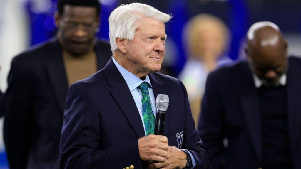 An older man with white hair, wearing a dark suit, blue shirt, and green patterned tie, holds a microphone while speaking. Two men stand behind him with heads bowed, dressed in dark suits. The background is blurred, suggesting an indoor event or ceremony.