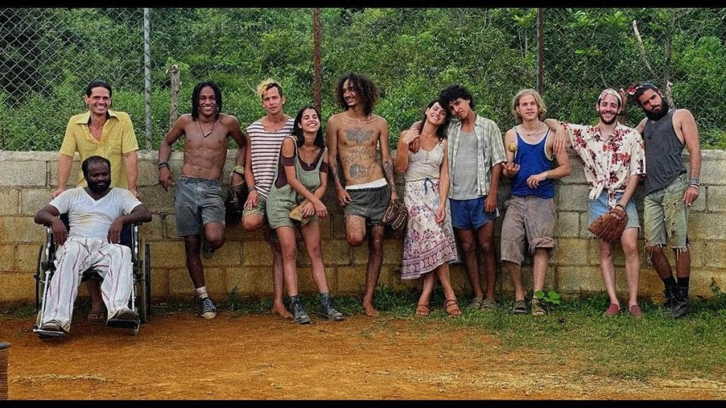 Ten people pose outdoors against a low brick wall and chain-link fence, with greenery behind. They wear casual, mismatched summer clothes. One man is in a wheelchair. The group appears relaxed and cheerful, some smiling, standing closely or with arms around each other.