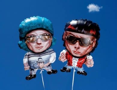 Two cartoon-style balloons float against a clear blue sky with a small white cloud. The left balloon shows a person with blue hair, sunglasses, and a blue outfit. The right balloon depicts a person with black hair, red scarf, and sunglasses. Both have exaggerated features.