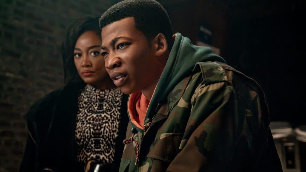A young man in a camouflage jacket and orange hoodie looks intensely ahead, while a woman in a black fur coat and patterned dress stands behind him, watching. Both have serious expressions and are in a dimly lit indoor setting with a brick wall background.