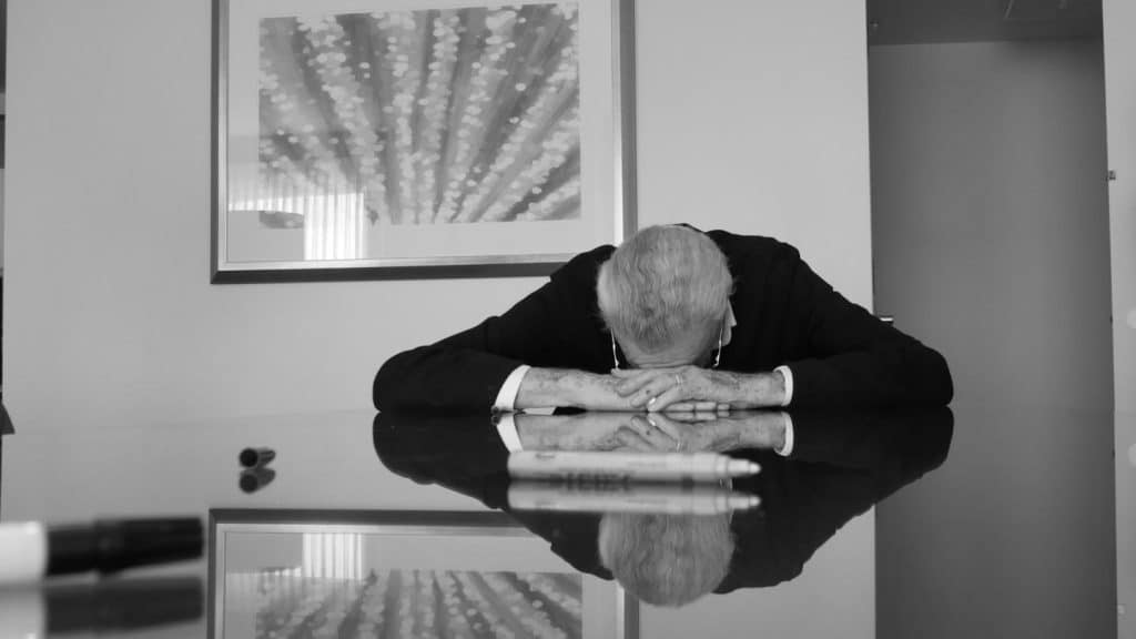 A person in a suit sits at a reflective table with their head resting on folded arms, facing down. Framed abstract art hangs on the wall behind them. A marker and pen lie on the table in the foreground, and the scene appears somber or contemplative.