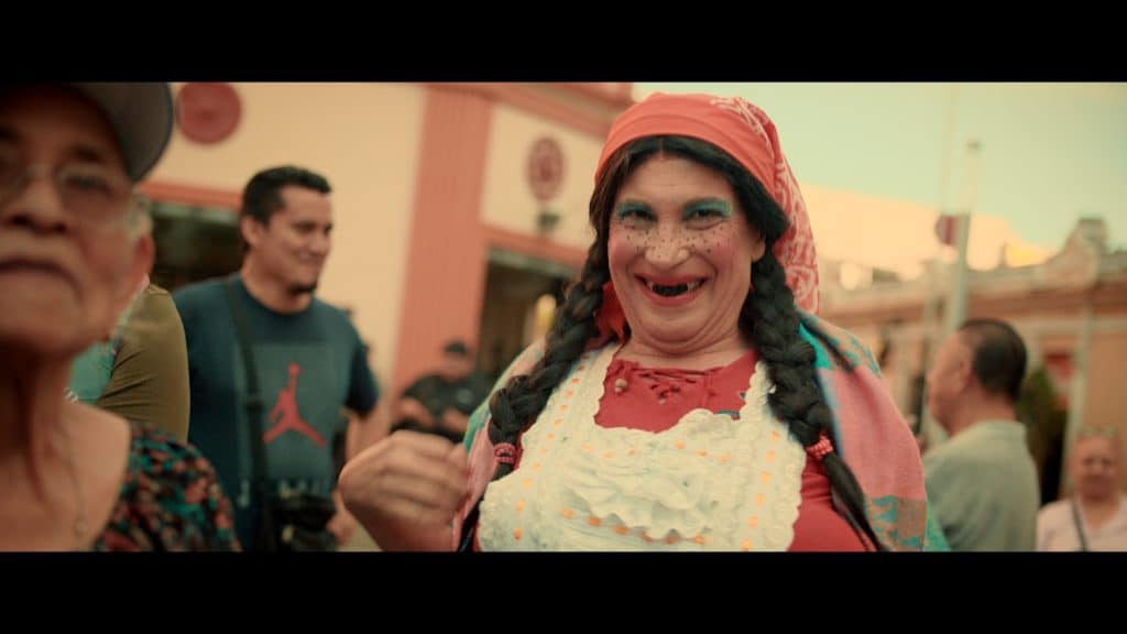 A smiling person in costume with braided hair, bright makeup, red headscarf, and apron stands in a festive outdoor crowd at an event celebrating La Balada de Hortensia, El Salvador's highest grossing comedy film. The joyful atmosphere is unmistakable.
