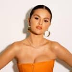 A woman with slicked-back dark hair wears large gold hoop earrings, a delicate necklace, and a strapless orange corset top. She poses confidently against a plain white background with her arms slightly out to the sides.
