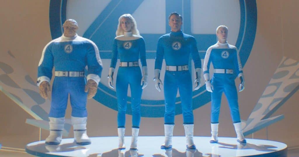 Four people in blue superhero suits with white gloves, belts, and boots stand side by side on a lit platform. Each suit features a “4” on the chest. The background is bright with a large blue circle and “4” insignia behind them.