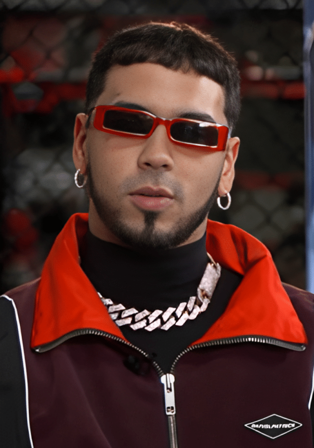 Anuel AA, A man with short dark hair and a neatly trimmed goatee wears red rectangular sunglasses, silver hoop earrings, a chunky silver chain, and a burgundy jacket with a red collar—ready for the Real Hasta La Muerte 2 Tour. The background is blurred fencing and red ropes.