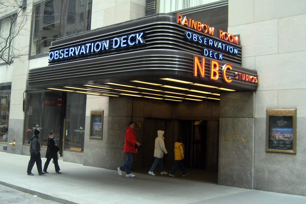 People walk past the entrance to Rockefeller Center in New York City, featuring an Art Deco marquee with neon signs for NBC Studios—iconic even amid recent NBC show cancellations 2025. The sidewalk and stone facade are visible.