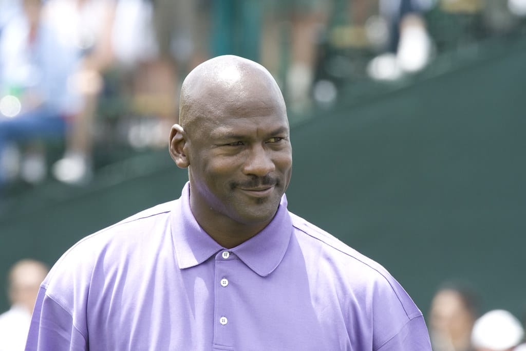 A man with a bald head and mustache stands outdoors in a lavender polo, slightly smiling. Blurred people and green stadium seating hint at NBA on NBC coverage 2025 in the background, with sunny lighting completing the scene.