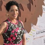 A woman with short curly hair smiles, wearing a sleeveless black dress adorned with pink and white floral embroidery. She stands in front of a backdrop featuring brown abstract shapes and oversized ticket stubs with “THE BEAR” and “FX Hulu” text visible.