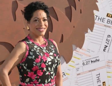 A woman with short curly hair smiles, wearing a sleeveless black dress adorned with pink and white floral embroidery. She stands in front of a backdrop featuring brown abstract shapes and oversized ticket stubs with “THE BEAR” and “FX Hulu” text visible.