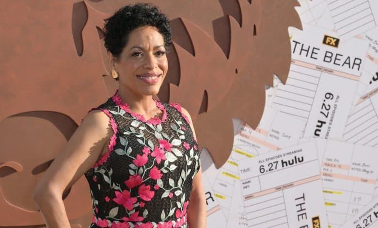 A woman with short curly hair smiles, wearing a sleeveless black dress adorned with pink and white floral embroidery. She stands in front of a backdrop featuring brown abstract shapes and oversized ticket stubs with “THE BEAR” and “FX Hulu” text visible.