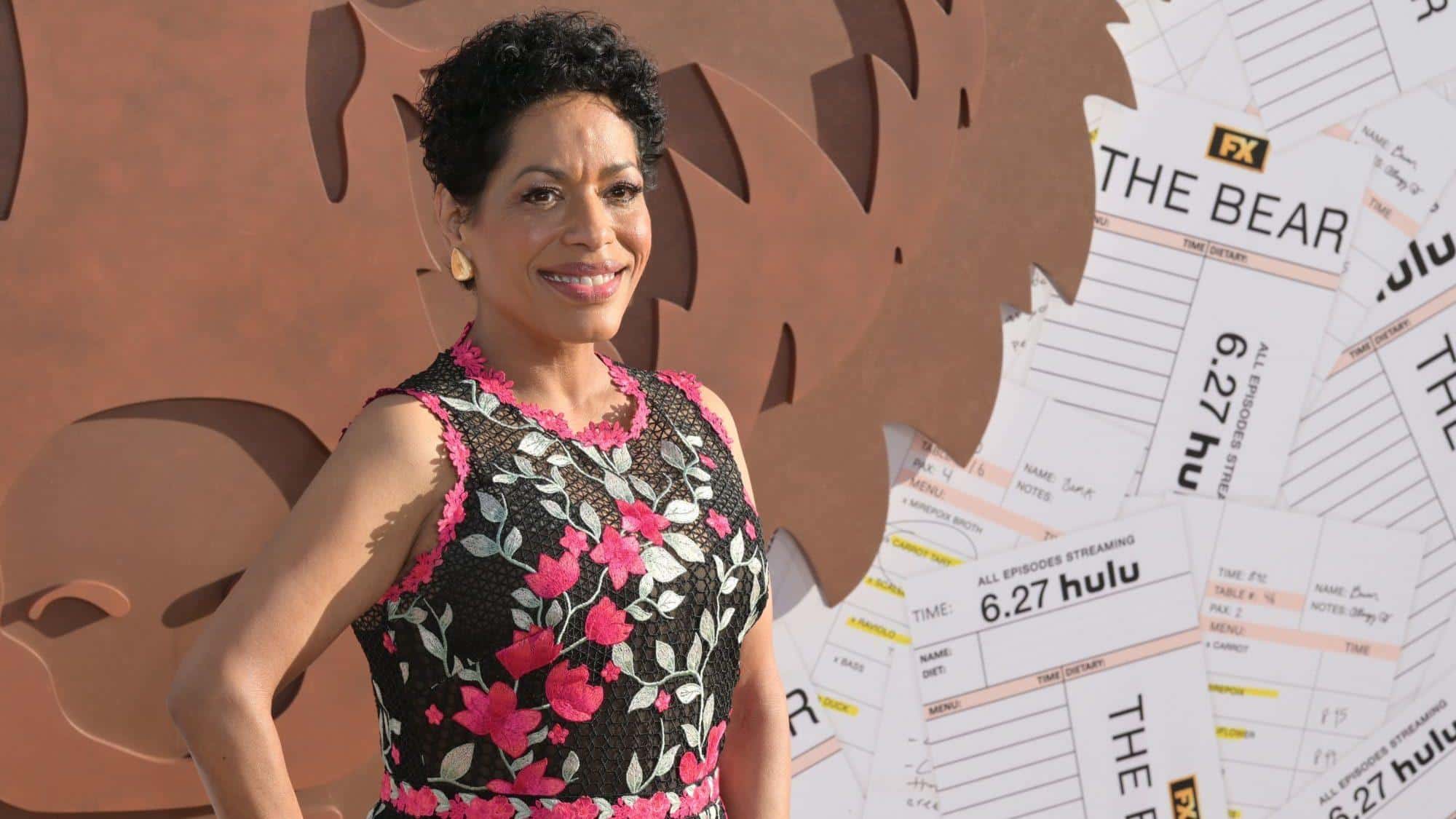 A woman with short curly hair smiles, wearing a sleeveless black dress adorned with pink and white floral embroidery. She stands in front of a backdrop featuring brown abstract shapes and oversized ticket stubs with “THE BEAR” and “FX Hulu” text visible.