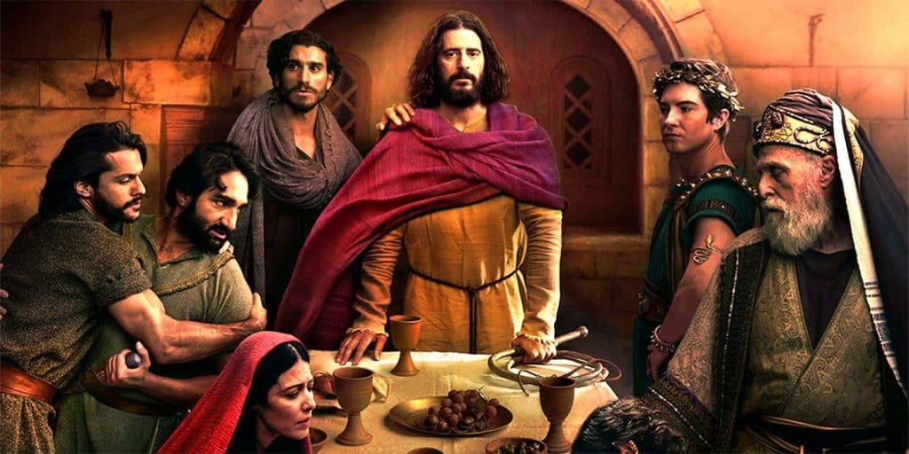 A group of people in historical robes gather around a table with bread, grapes, and goblets in a stone room. One central figure in yellow and red stands out, flanked by others with somber expressions, suggesting a dramatic or biblical scene.