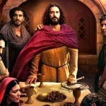 A group of people in historical robes gather around a table with bread, grapes, and goblets in a stone room. One central figure in yellow and red stands out, flanked by others with somber expressions, suggesting a dramatic or biblical scene.