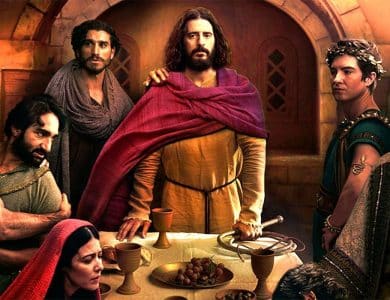 A group of people in historical robes gather around a table with bread, grapes, and goblets in a stone room. One central figure in yellow and red stands out, flanked by others with somber expressions, suggesting a dramatic or biblical scene.