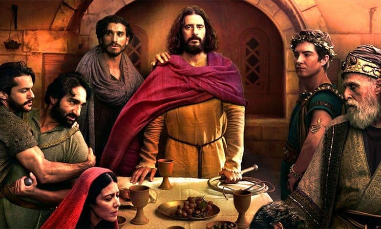 A group of people in historical robes gather around a table with bread, grapes, and goblets in a stone room. One central figure in yellow and red stands out, flanked by others with somber expressions, suggesting a dramatic or biblical scene.