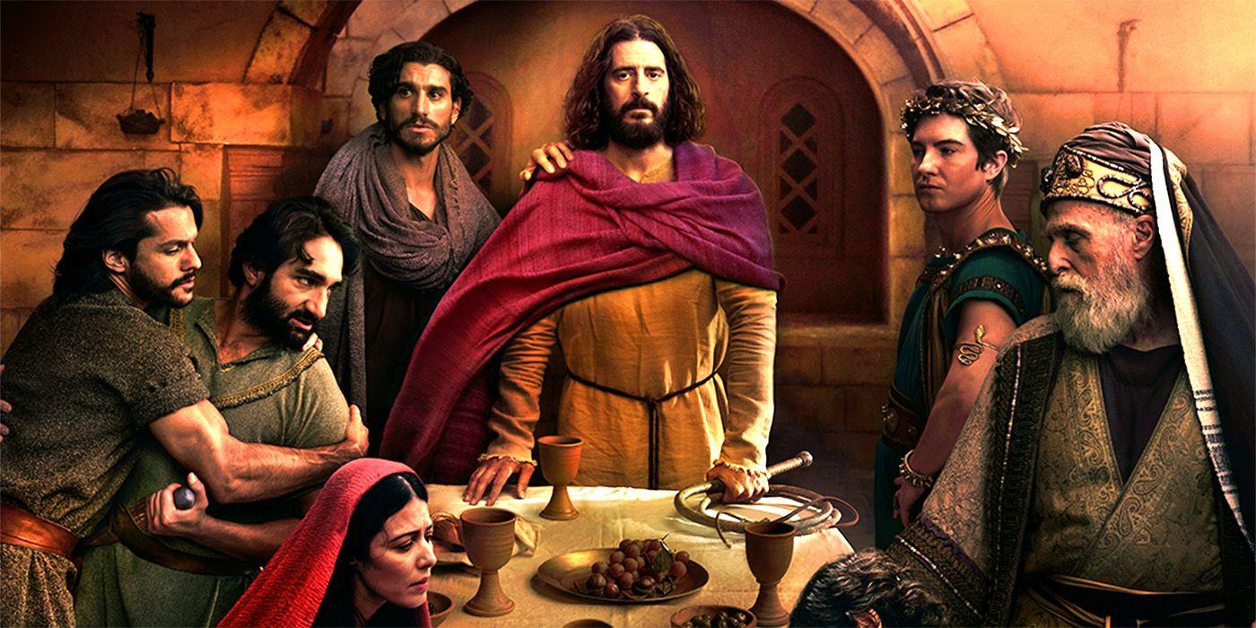 A group of people in historical robes gather around a table with bread, grapes, and goblets in a stone room. One central figure in yellow and red stands out, flanked by others with somber expressions, suggesting a dramatic or biblical scene.