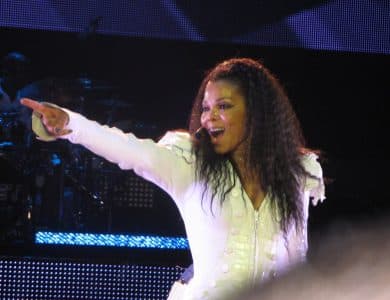 A woman in a white, detailed jacket smiles and points energetically while performing on stage, her long, curly hair framing her face. With the stage lights behind her, she radiates Janet Jackson ICON energy before the shadowy drum set.