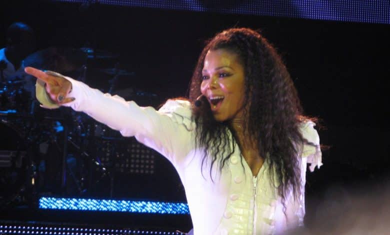A woman in a white, detailed jacket smiles and points energetically while performing on stage, her long, curly hair framing her face. With the stage lights behind her, she radiates Janet Jackson ICON energy before the shadowy drum set.