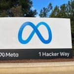 A large sign with the blue Meta logo, resembling an infinity symbol, stands above "Meta" and "1 Hacker Way." The Meta sign is outdoors, framed by green trees and a blue sky dotted with clouds.