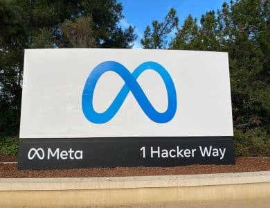A large sign with the blue Meta logo, resembling an infinity symbol, stands above "Meta" and "1 Hacker Way." The Meta sign is outdoors, framed by green trees and a blue sky dotted with clouds.