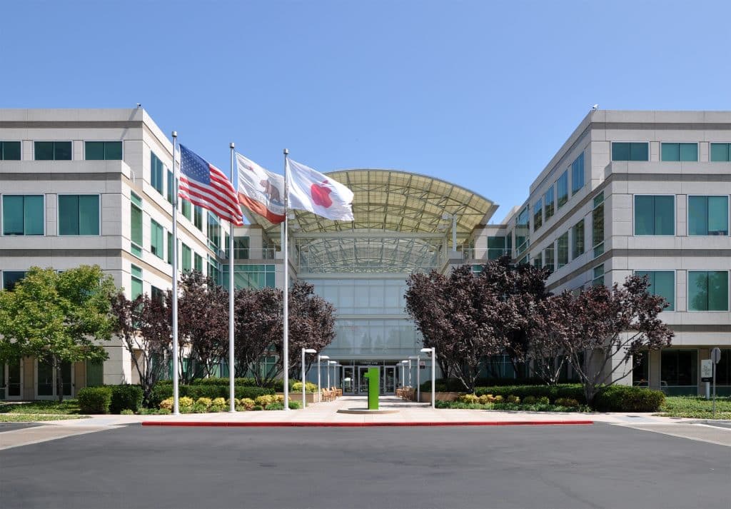 Apple Headquarters in Cupertino, A modern office building with a glass entrance, light-colored facade, and symmetrical design is shown. Three flags—the American flag, a white flag, and a California state flag—are flying in front. Trees and bushes line the walkway leading to the entrance.